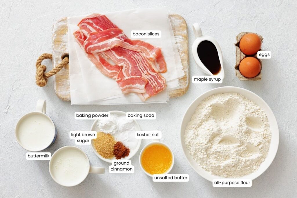 Assorted labeled ingredients on a white surface, including bacon slices, eggs, flour, buttermilk, brown sugar, cinnamon, butter, baking powder, baking soda, salt, and maple syrup.
