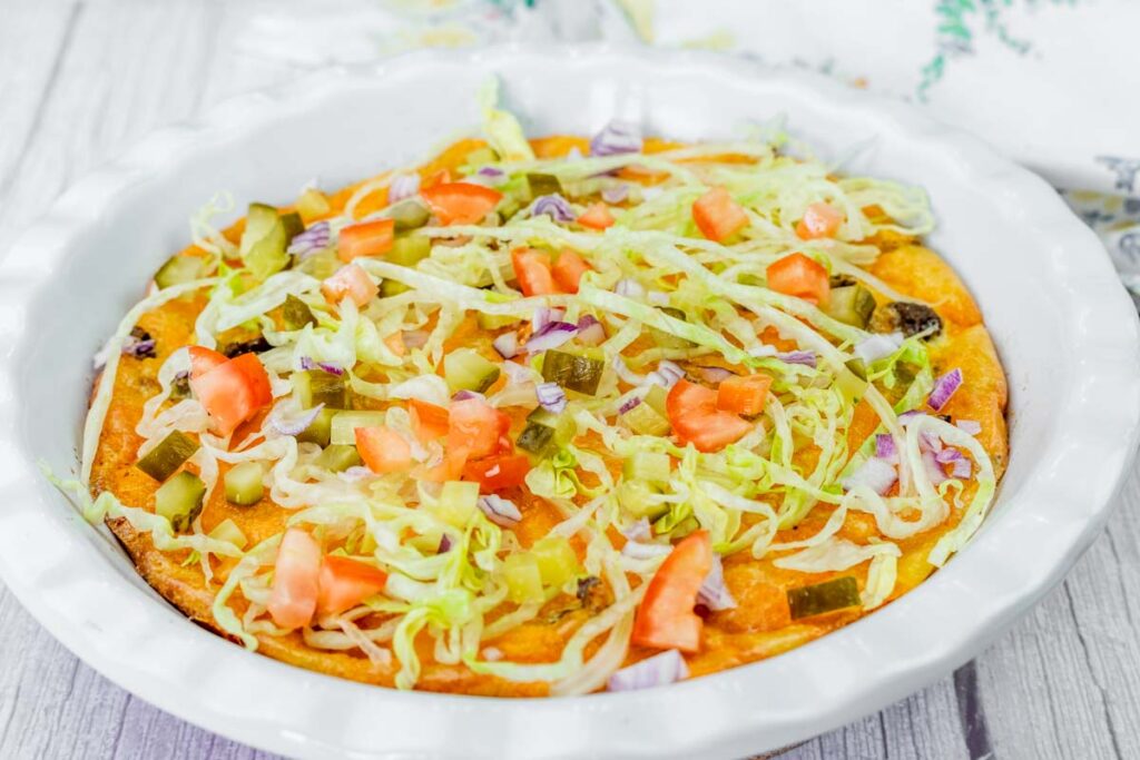 A baked dish topped with shredded lettuce, diced tomatoes, pickles, and chopped red onions in a white round baking dish on a light surface.
