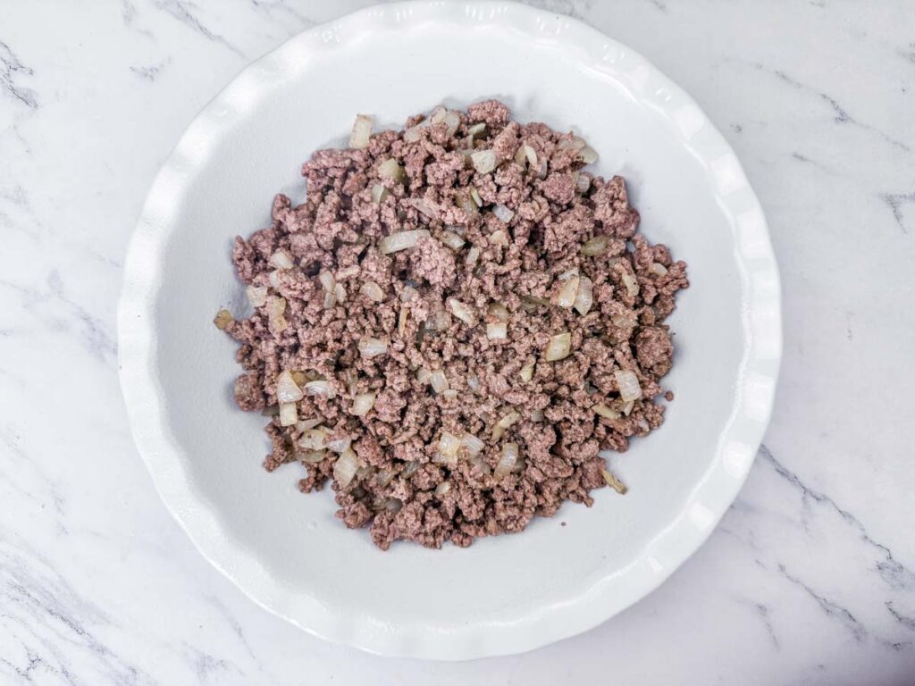A white plate with cooked ground beef and chopped onions sits on a marble surface.