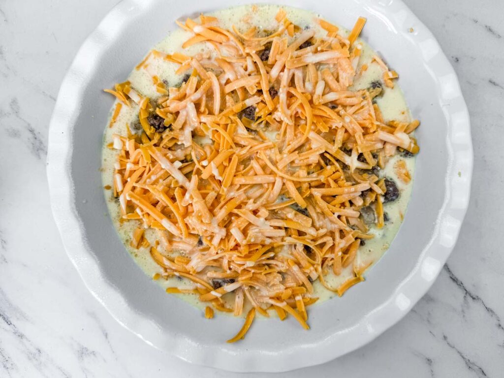 A pie dish filled with an uncooked mixture of beaten eggs, mushrooms, and shredded cheddar cheese on a marble surface.