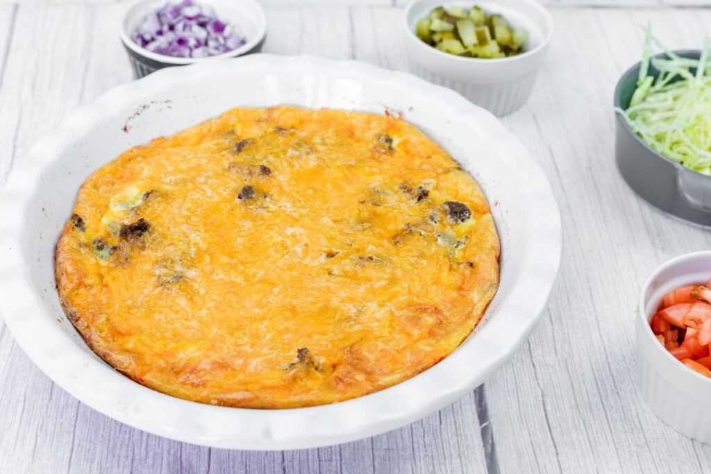 A baked egg casserole with a golden cheese top in a white dish, surrounded by small bowls of diced onion, pickles, shredded lettuce, and chopped tomato.