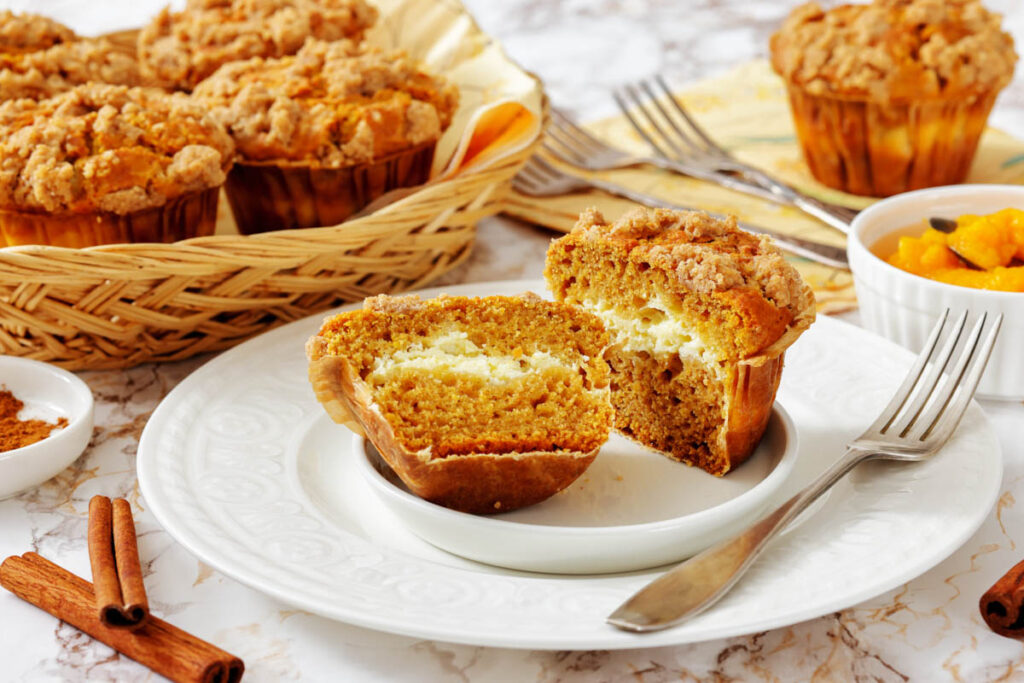 A pumpkin streusel muffin cut in half on a white plate, showing a cream cheese filling, with more muffins and cinnamon sticks in the background.