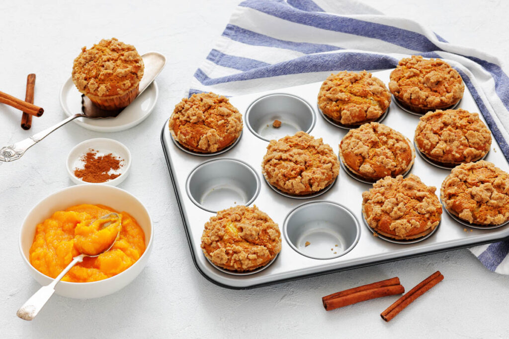A muffin tray with pumpkin streusel muffins, a bowl of pumpkin puree, cinnamon sticks, and a striped towel on a white surface. One muffin is served on a plate.