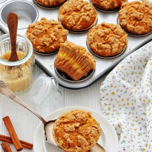 A tray of baked pumpkin muffins with crumb topping, one muffin on a plate with a spoon, a jar of brown sugar, cinnamon sticks, and a floral cloth nearby.