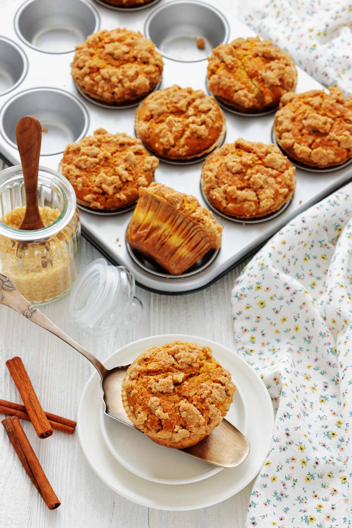 A tray of baked pumpkin muffins with crumb topping, one muffin on a plate with a spoon, a jar of brown sugar, cinnamon sticks, and a floral cloth nearby.