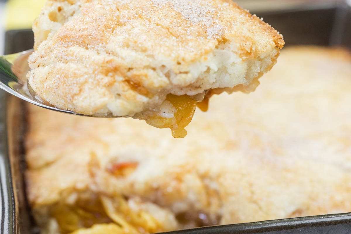 A close-up of a serving of peach cobbler being lifted from a baking dish, showing a golden, flaky crust with visible peach filling.