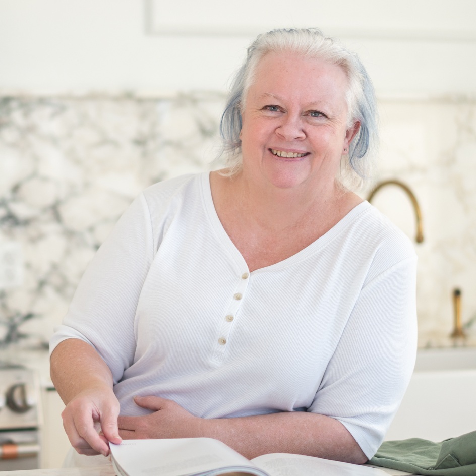 An older woman with light gray hair and a white shirt sits at a kitchen counter, smiling and holding open a book.