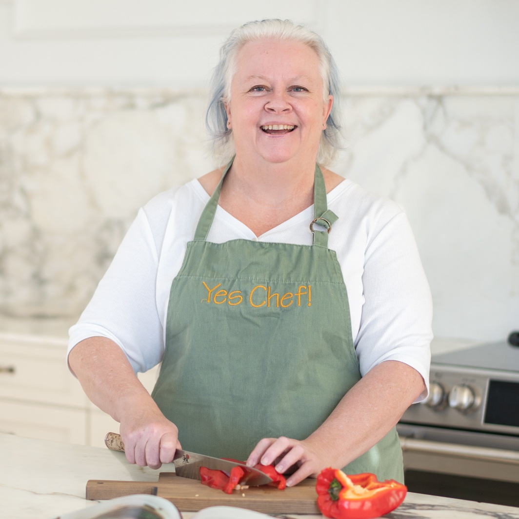 Chef Jenn wearing a green "Yes Chef!" apron smiles while slicing a red bell pepper on a cutting board in a kitchen.