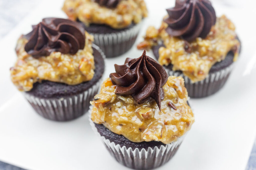 Four chocolate cupcakes topped with a layer of coconut-pecan frosting and a swirl of chocolate frosting, arranged on a white plate.