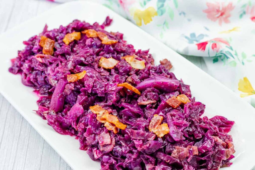 A rectangular white plate with cooked red cabbage and pieces of bacon displayed on a light wooden surface next to a floral-patterned cloth.