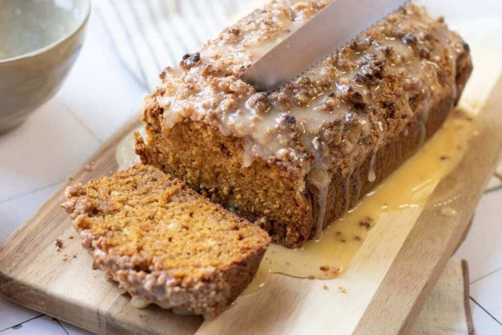 A loaf of pumpkin bread with streusel sits on a wooden cutting board; one slice is cut, and a knife rests on top.