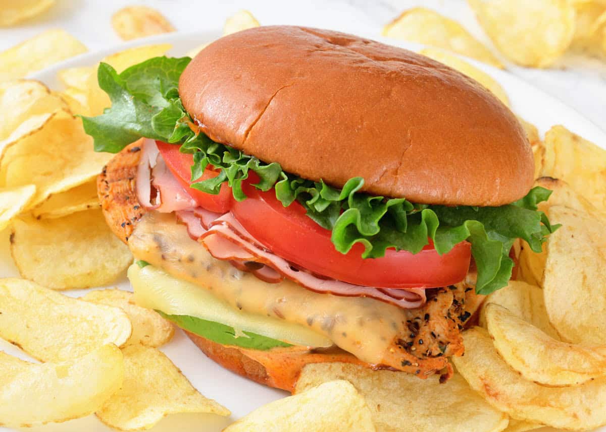 A grilled California chicken burger with lettuce, tomato, cheese, and ham on a bun, served with potato chips on a white plate.