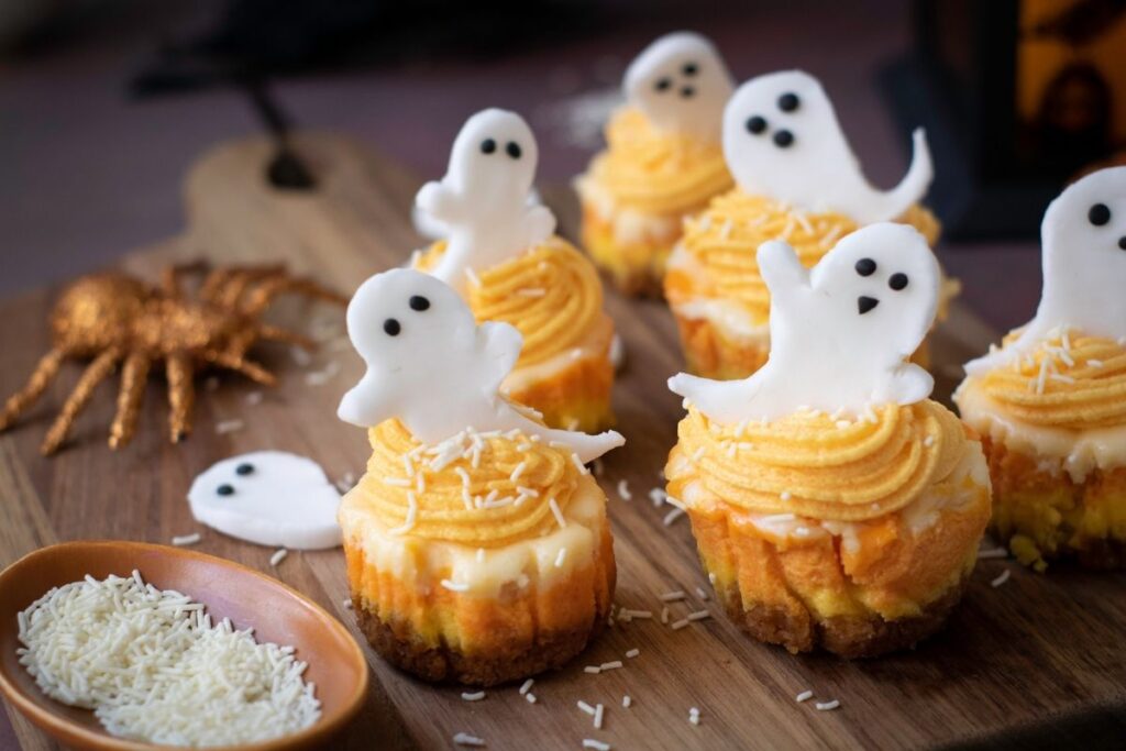 Cupcakes with yellow frosting, white sprinkles on top, and white fondant ghost decorations are arranged on a wooden board, with a spider decoration and a bowl of white sprinkle nearby.