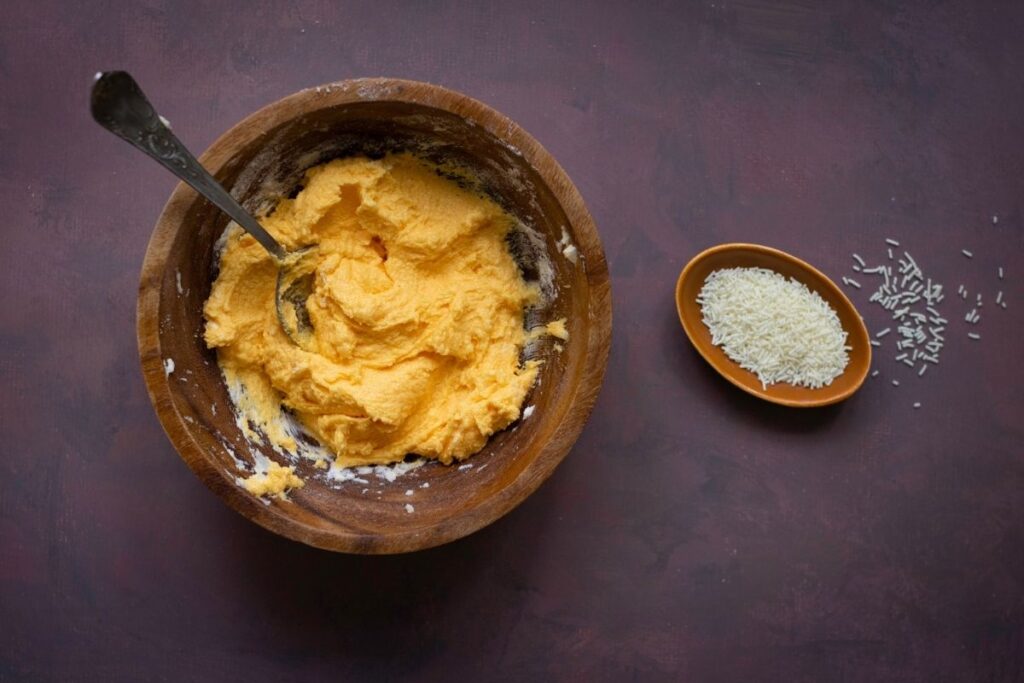 A wooden bowl with a spoon contains a yellow frosting mixture. Nearby, a small dish holds white sprinkles with some scattered on the table.