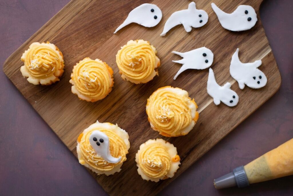 A wooden board holds six frosted cupcakes and several small white fondant ghost decorations, with a piping bag nearby.