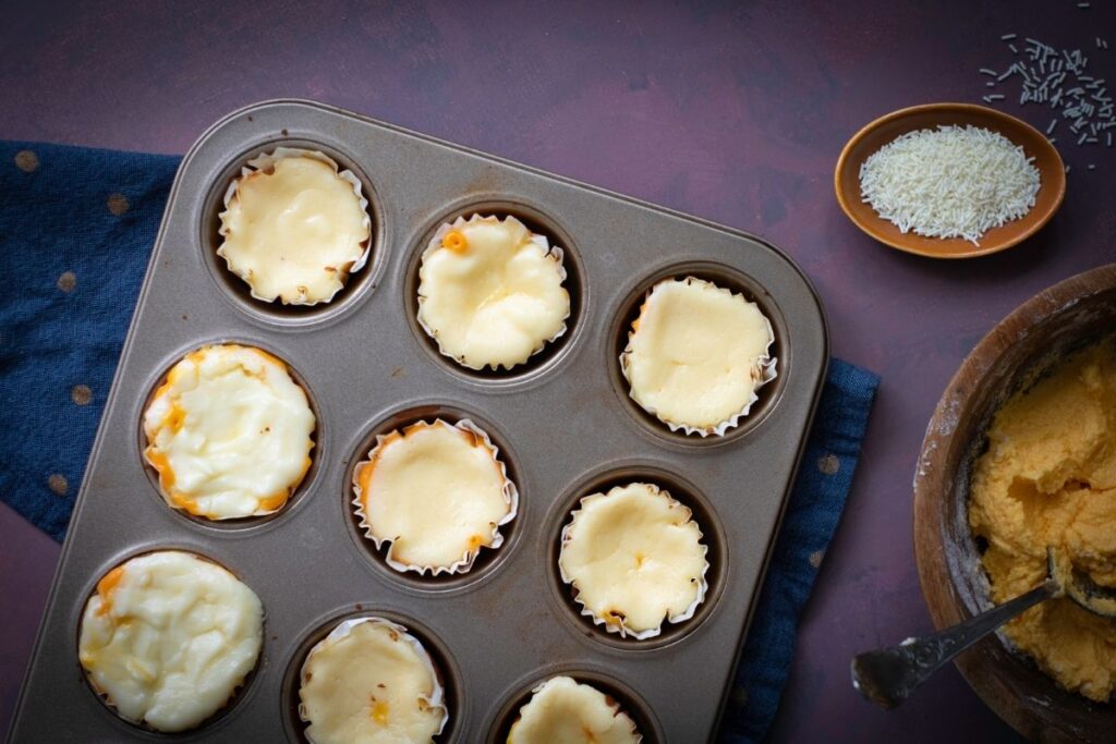 Baked cupcakes in a muffin tin, with a bowl of white sprinkles and a wooden bowl of orange frosting nearby.