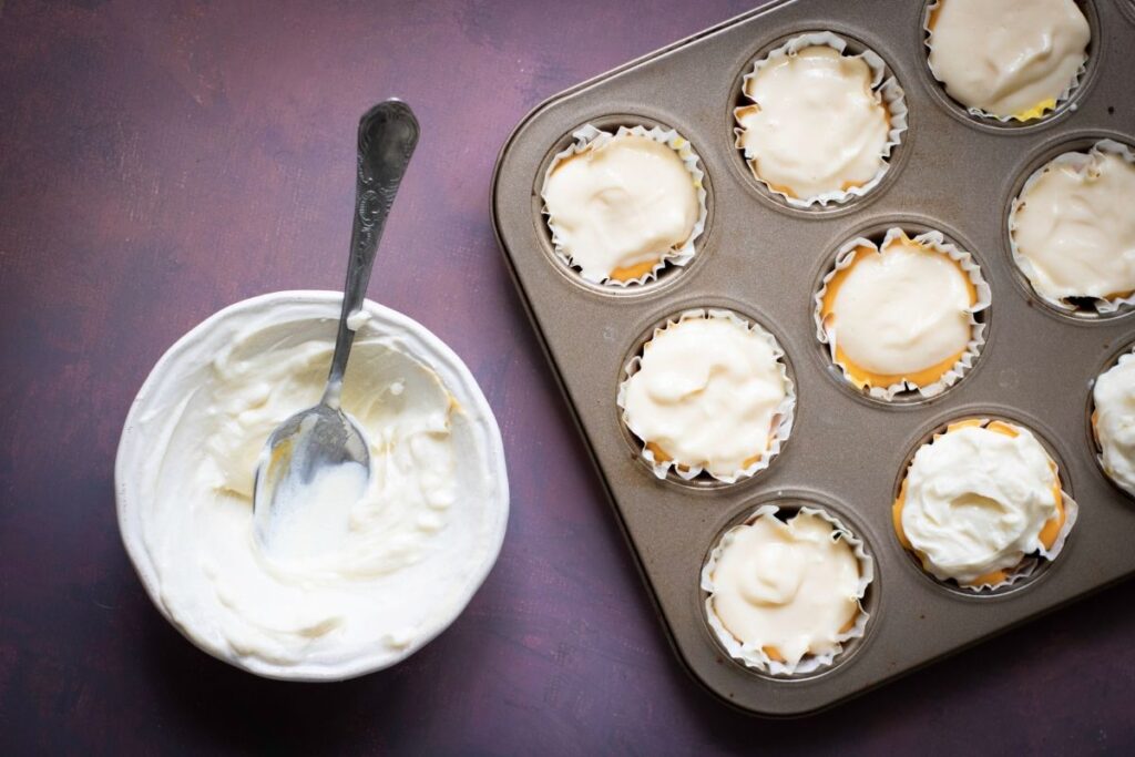 A muffin tin with cupcakes topped with batter next to a bowl of batter and a spoon on a dark surface.