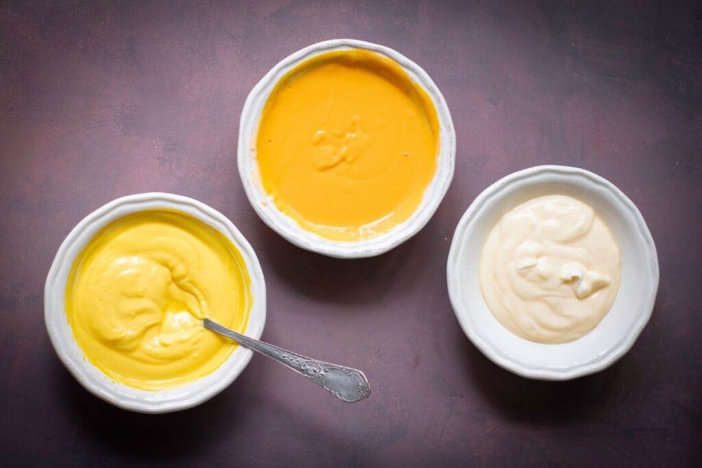 Three white bowls filled with different creamy batter—yellow, orange, and off-white—are arranged on a dark surface, with a spoon in the yellow batter.