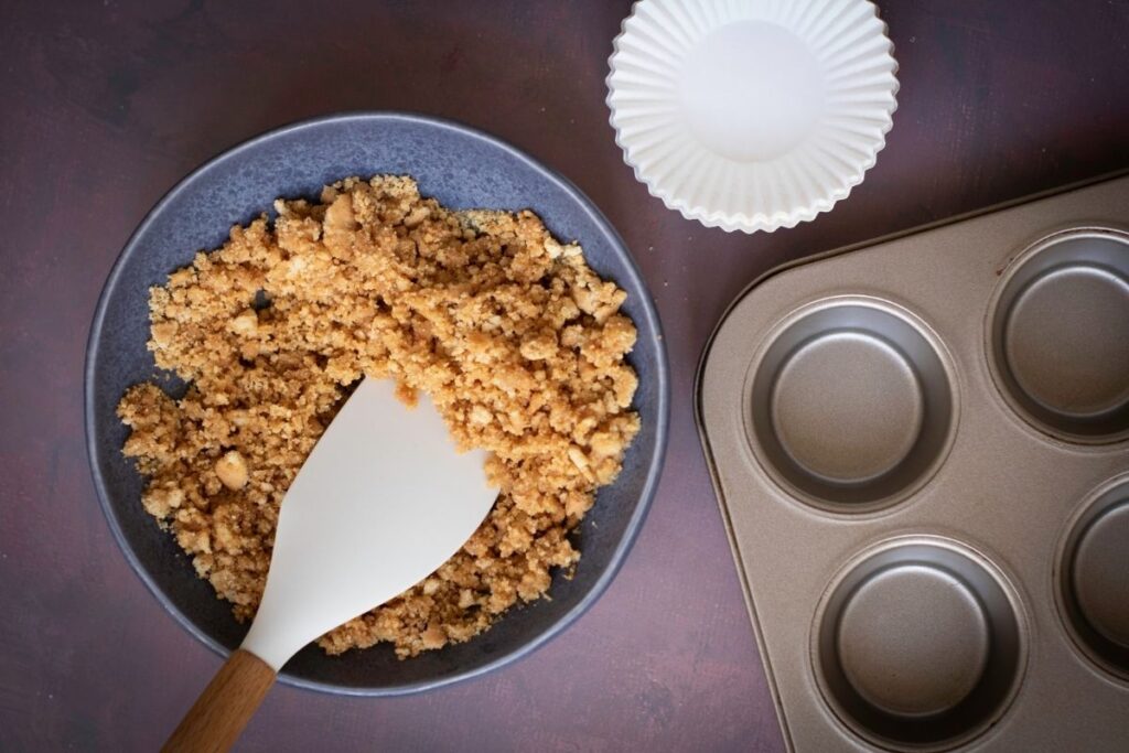 A mixture of graham cracker crumbs, sugar, and melted butter in a bowl with a spatula. Nearby are a muffin tin and paper cupcake liners.