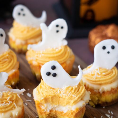 Cupcakes with yellow frosting and white fondant ghost decorations are arranged on a wooden board, with white sprinkles on top.