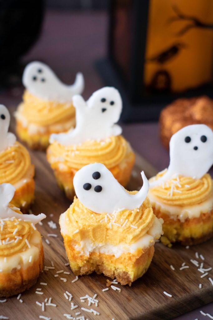 Cupcakes with yellow frosting and white fondant ghost decorations are arranged on a wooden board, with white sprinkles on top.