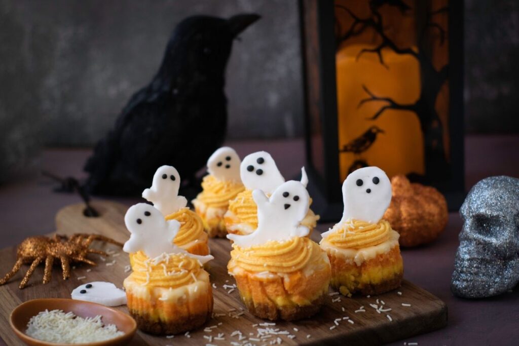 Cupcakes with yellow frosting and white fondant ghost decorations are arranged on a wooden board, with white sprinkles on top. In the background are Halloween decorations such as a lantern, skull, spider, and crow.