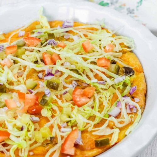 A baked dish topped with shredded lettuce, diced tomatoes, chopped onions, pickles, and melted cheese in a white ceramic pie dish.
