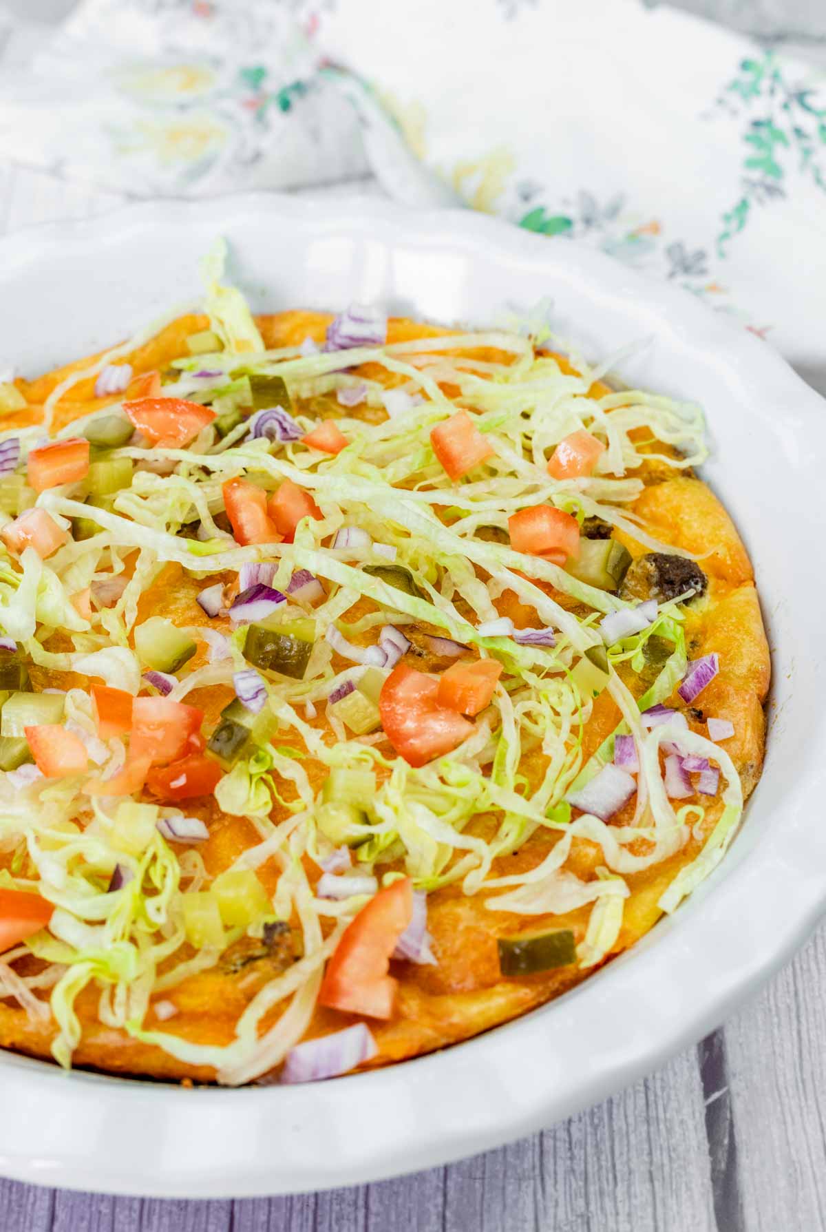 A baked dish topped with shredded lettuce, diced tomatoes, chopped onions, pickles, and melted cheese in a white ceramic pie dish.