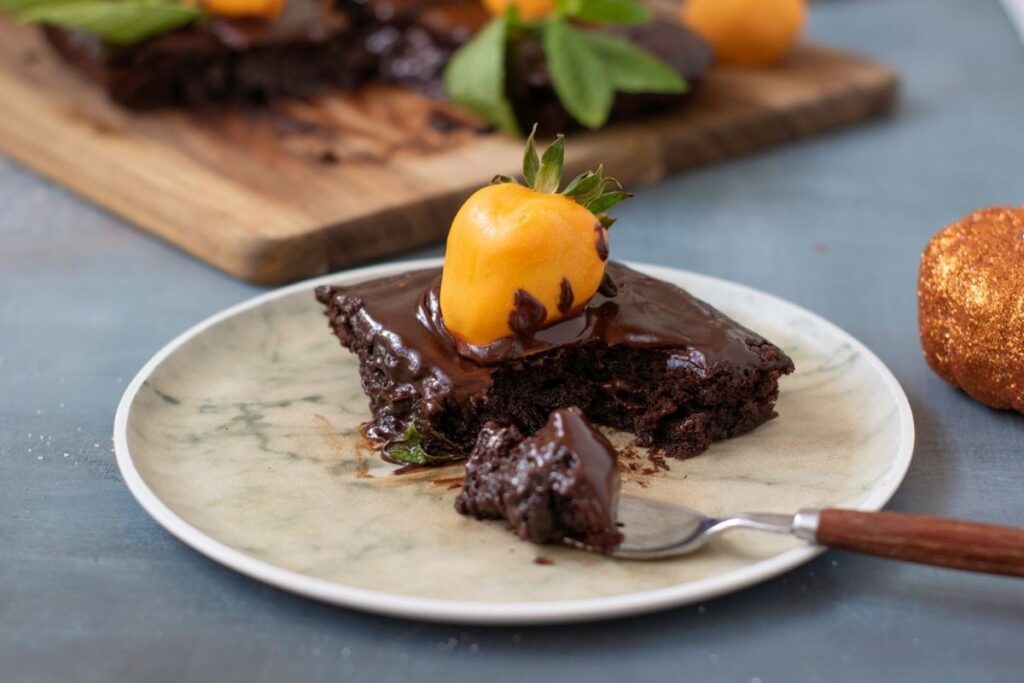 A piece of Pumpkin Patch Brownie Cake sits on a plate with a fork. More cake is visible in the background on a wooden board.