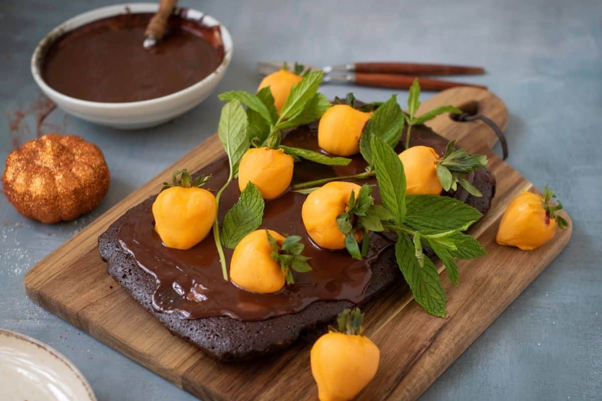 A rectangular Halloween Pumpkin Patch Brownie Cake, topped with yellow fruit resembling mini pumpkins and fresh mint, sits on a wooden board.