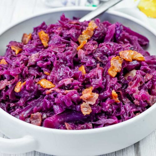A white bowl filled with cooked red cabbage and pieces of bacon, set on a light wooden surface.