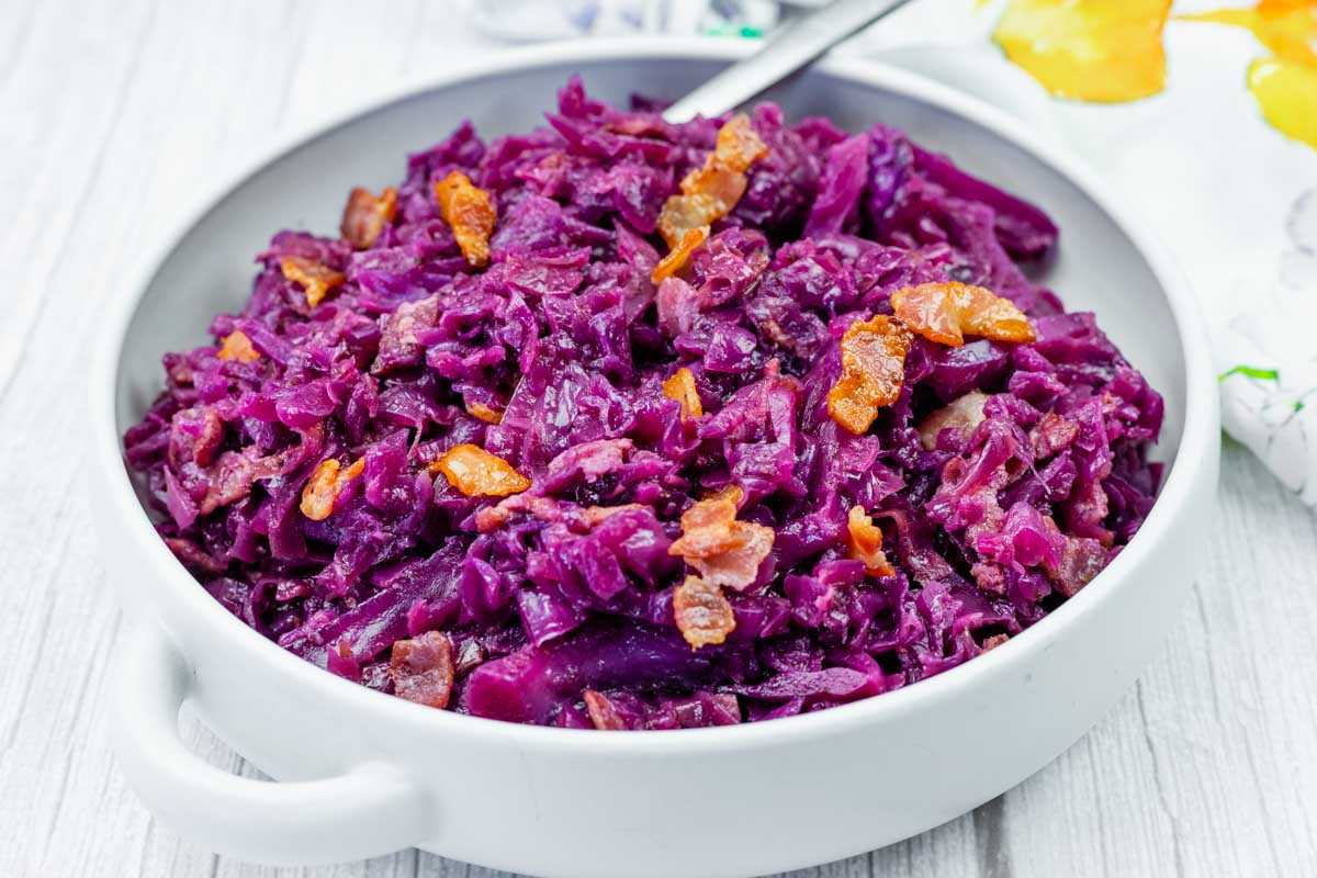 A white bowl filled with cooked red cabbage and pieces of bacon, set on a light wooden surface.