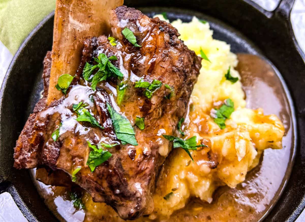 Braised beef short rib with gravy served over mashed potatoes, garnished with chopped fresh herbs in a black dish.