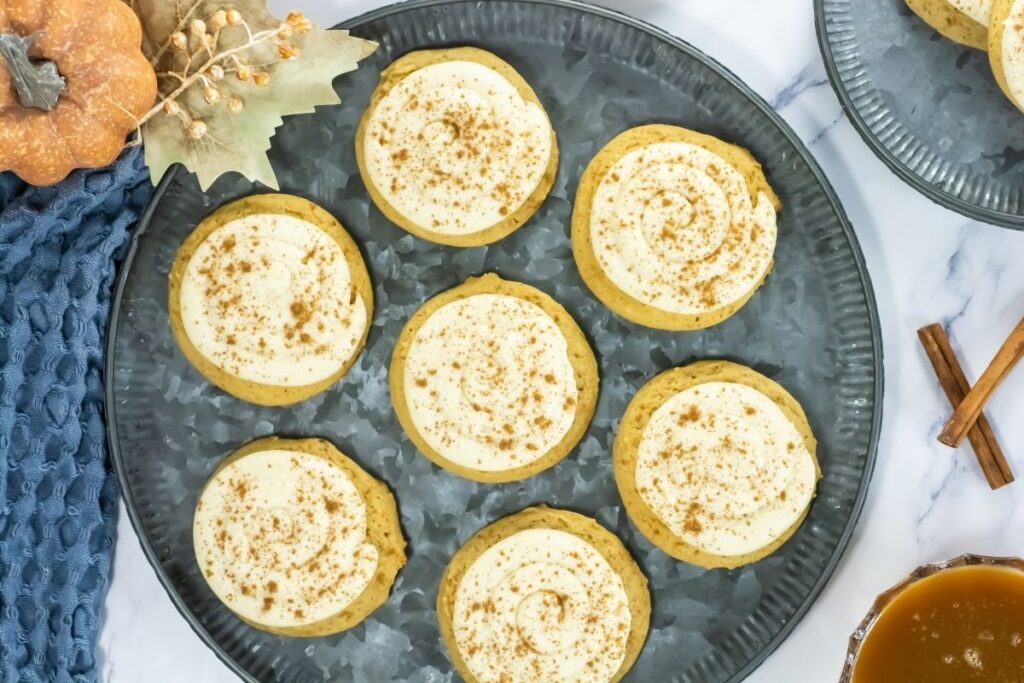 Crumbl Caramel-Pumpkin Cookies are arranged on a metal tray, accompanied by fall-themed decorations and cinnamon sticks.