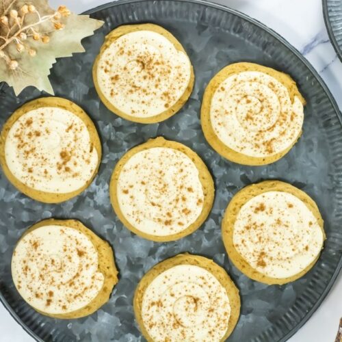Crumbl Caramel-Pumpkin Cookies are arranged on a metal tray, accompanied by fall-themed decorations and cinnamon sticks.