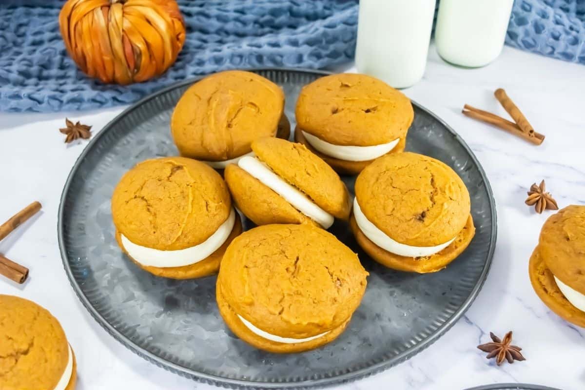 A plate with pumpkin whoopie pies filled with white cream, surrounded by cinnamon sticks, star anise, and a small pumpkin decoration.