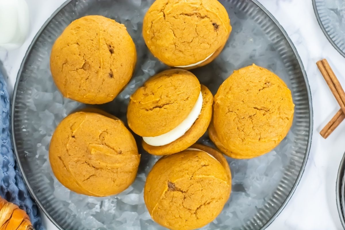 Six pumpkin whoopie pies with cream filling are arranged on a gray plate, viewed from above. Two cinnamon sticks are visible at the edge of the plate.