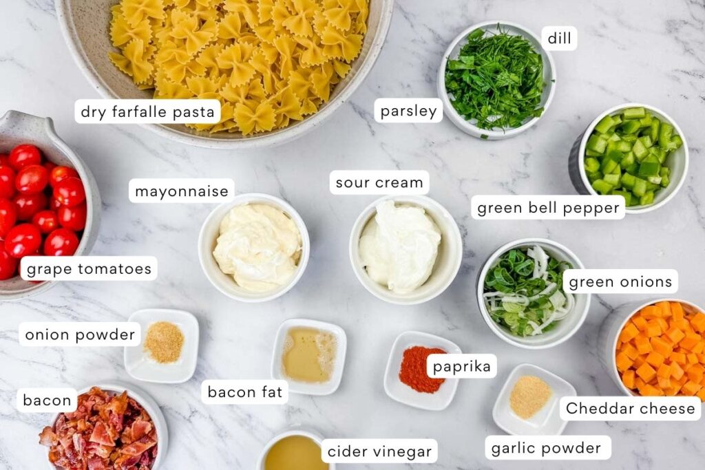 Dry farfalle pasta, mayonnaise, sour cream, grape tomatoes, green onions, green bell pepper, cheddar cheese, bacon, herbs, spices, and seasonings arranged on a marble surface.