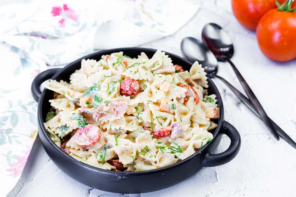 A black bowl filled with creamy bowtie pasta salad mixed with cherry tomatoes, herbs, and vegetables sits on a white surface next to two spoons and fresh tomatoes.