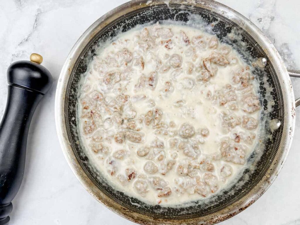 A skillet filled with sausage gravy sits on a marble countertop next to a black pepper grinder.