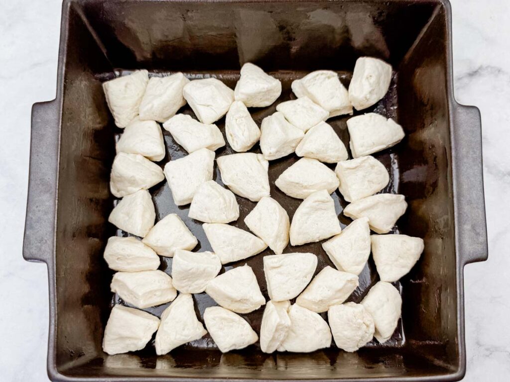 A square baking pan filled with evenly spaced, unbaked biscuit dough pieces on a light countertop.
