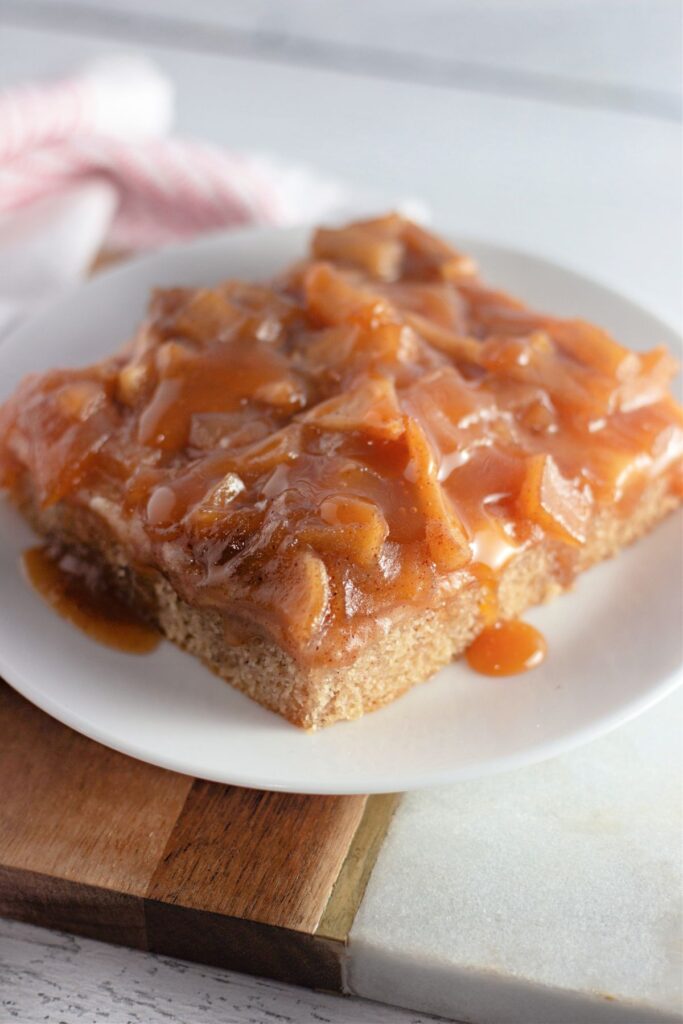 A square slice of cake topped with caramelized apple pieces and sauce sits on a white plate.
