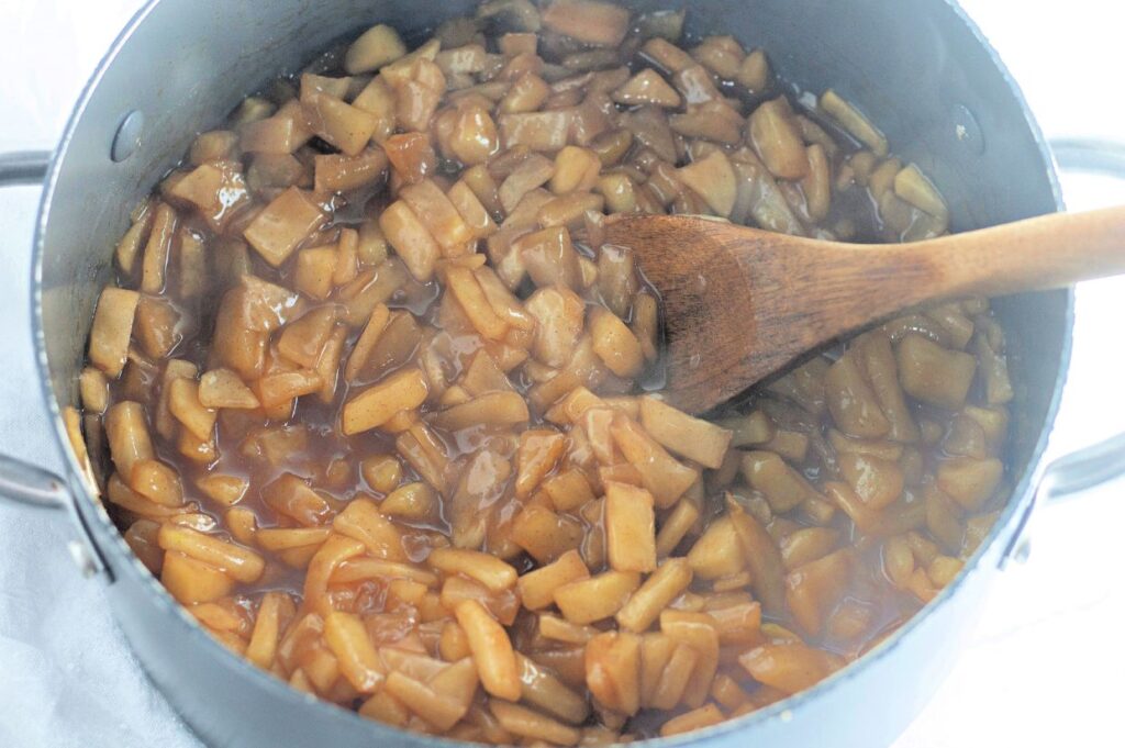 Diced apples cooking in a pot with brown syrup, stirred by a wooden spoon.