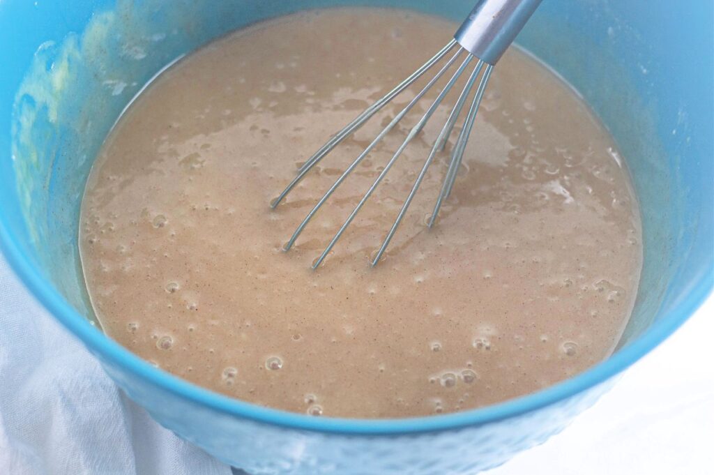 A metal whisk in a blue bowl mixing a light brown batter.