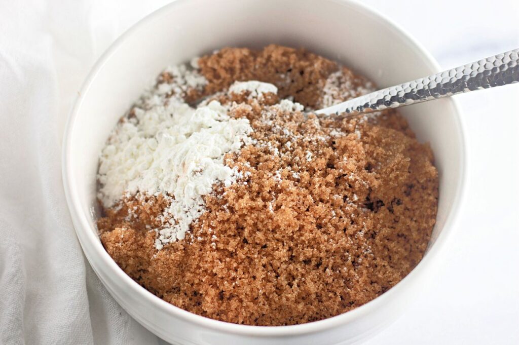 A white bowl filled with brown sugar and a small amount of white cornstarch, with a metal spoon resting inside.