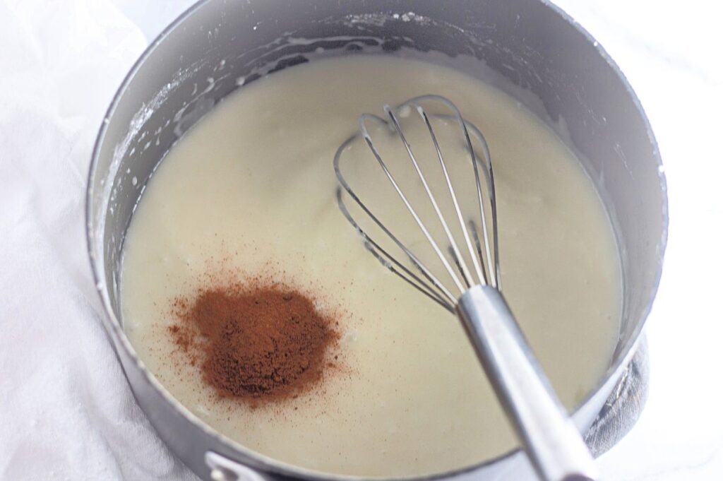 A metal whisk rests in a saucepan filled with a creamy white mixture and a heap of ground cinnamon on the surface.