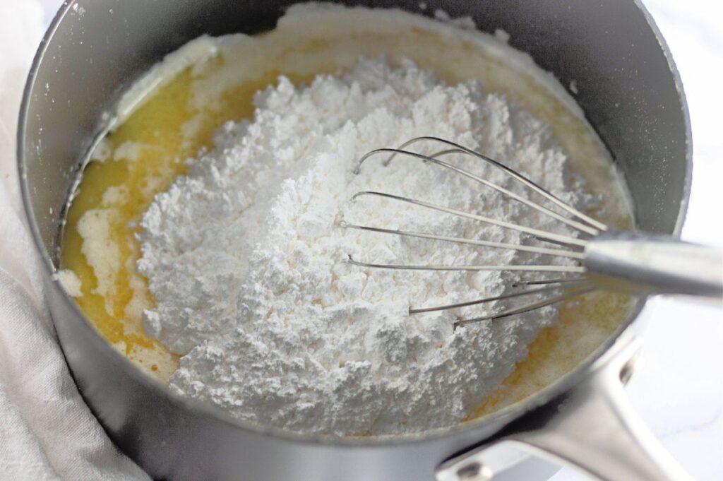 A whisk rests in a saucepan filled with melted butter and a mound of powdered sugar, ready to be mixed together.