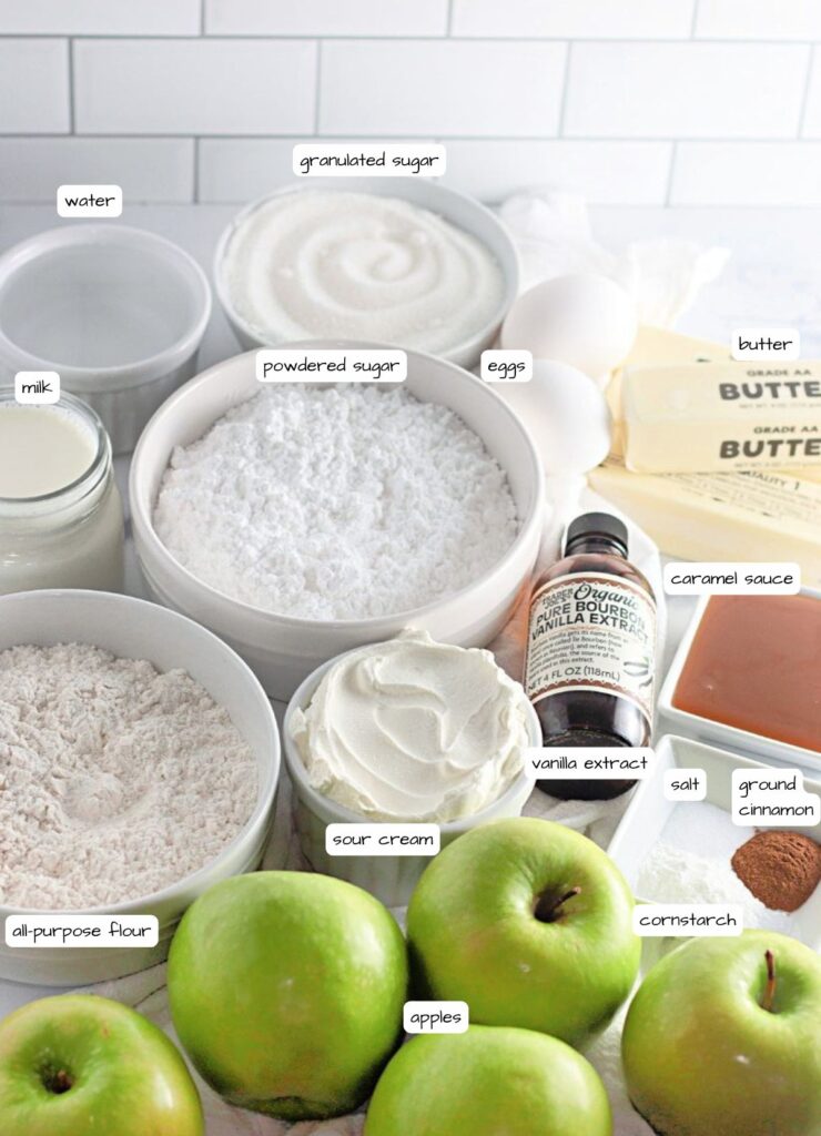 Ingredients for baking are arranged on a counter, including apples, flour, sugar, eggs, butter, milk, sour cream, vanilla extract, caramel sauce, cornstarch, salt, and cinnamon.
