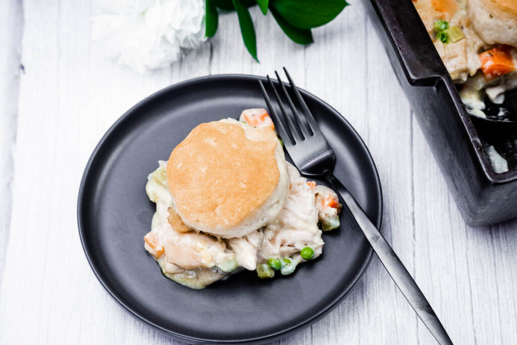 A plate with a serving of chicken and vegetable casserole topped with a biscuit, next to a fork; casserole dish is partially visible on the side.
