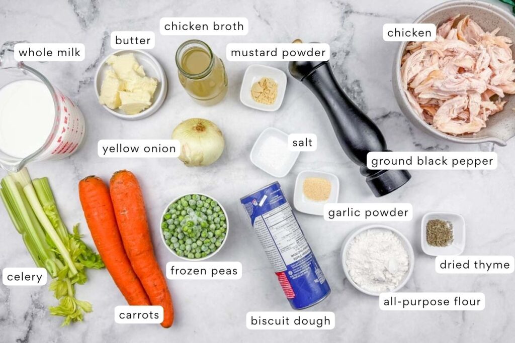 Ingredients for chicken pot pie laid out on a white surface, including chicken, vegetables, milk, butter, spices, chicken broth, frozen peas, and biscuit dough.
